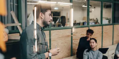 Skäggig man står och presenterar i ett möte framför två kollegor, en man och en kvinna. Bilden är tagen genom en fönsterruta och det syns en reflektion i bilden. Kontoret upplevs som en kreativ arbetsplats.