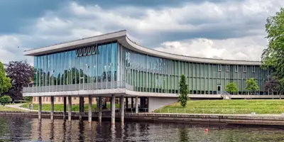  Halmstad universitets bibliotek med pelare i floden. Bild tagen med grönskande träd runtomkring.